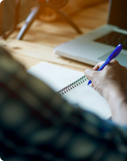 Aluno escreve em folha de caderno em frente a notebook